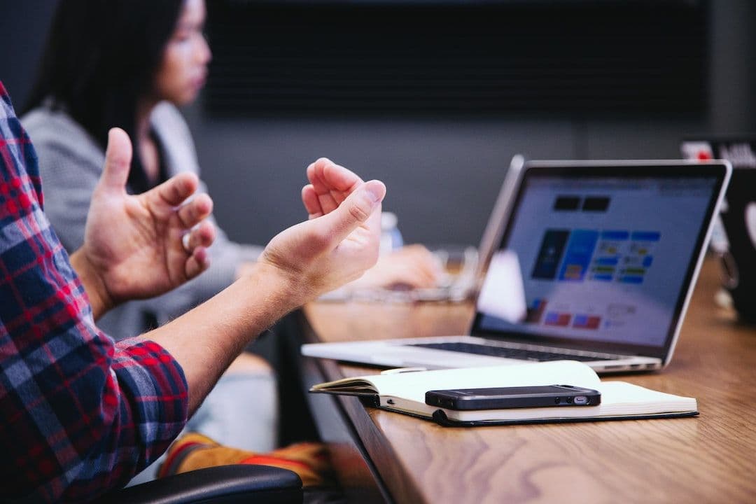A meeting table with a laptop, notebook and phone; a speaker’s hands in the foreground.